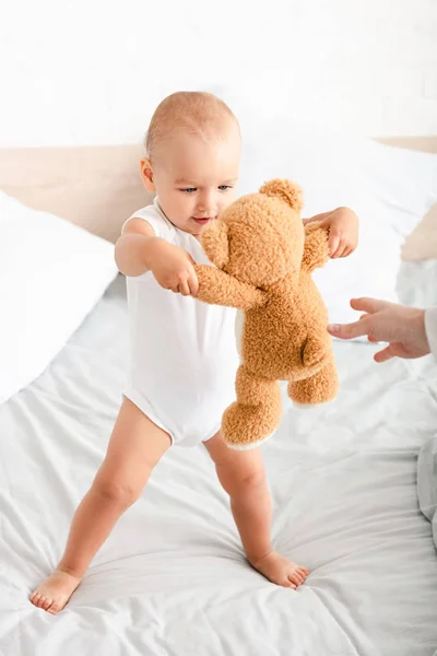 Niedliches Barfußkind in weißen Kleidern, das mit seinem Teddybär auf dem Bett spielt — Stockfoto