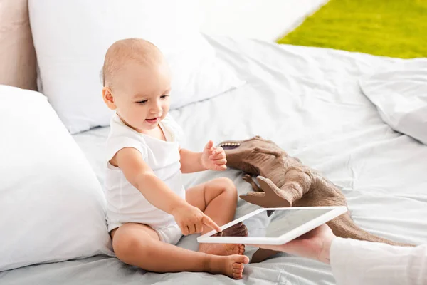 Lindo niño descalzo en ropa blanca apuntando con el dedo a la tableta digital cerca de dinosaurio juguete marrón - foto de stock