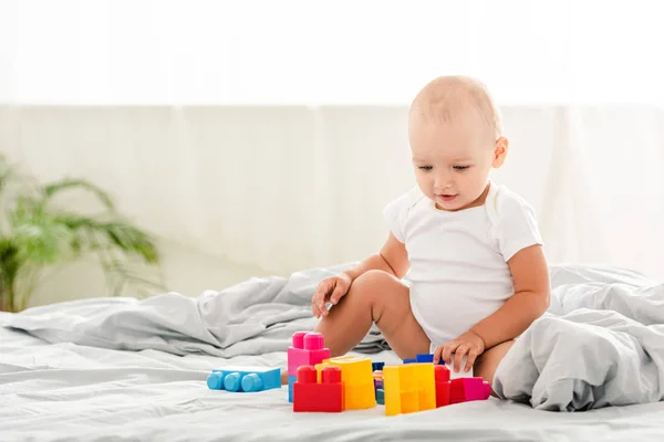 Bébé souriant en vêtements blancs assis sur le lit et regardant des jouets — Photo de stock