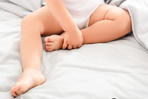 Обрізаний вид милої дитини, що сидить на ліжку і тримає ноги — стокове фото