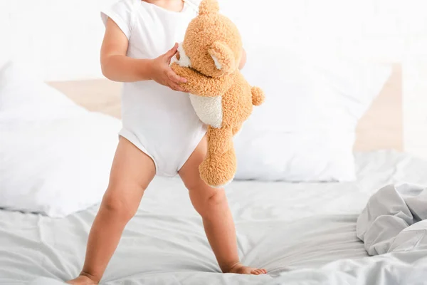 Bonito criança descalça em roupas brancas em pé na cama com seu urso de brinquedo marrom — Fotografia de Stock