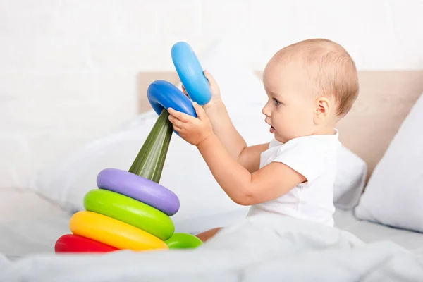 Vista lateral de lindo niño pequeño en ropa blanca jugando con la pirámide de juguete en la cama - foto de stock