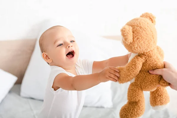 Симпатичный маленький ребенок в белой одежде открывает рот, улыбаясь и поднимая руки к коричневому плюшевому медведю — стоковое фото