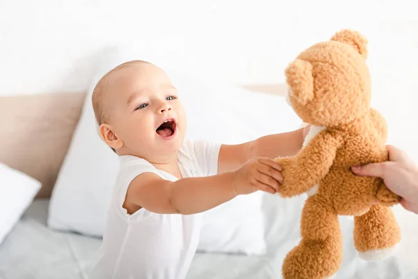 Lindo niño pequeño con ropa blanca abriendo la boca, sonriendo y levantando las manos para oso de peluche marrón - foto de stock