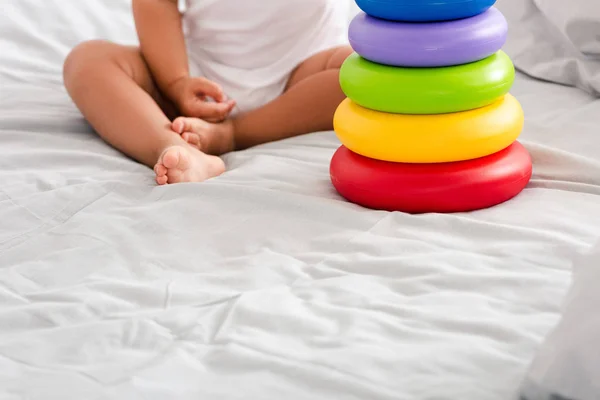 Vista parcial de un niño descalzo con ropa blanca sentado en la cama cerca de la pirámide de juguetes - foto de stock