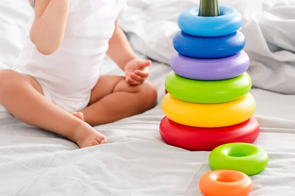 Vista recortada de un niño descalzo con ropa blanca sentado en la cama cerca de la pirámide de juguetes - foto de stock