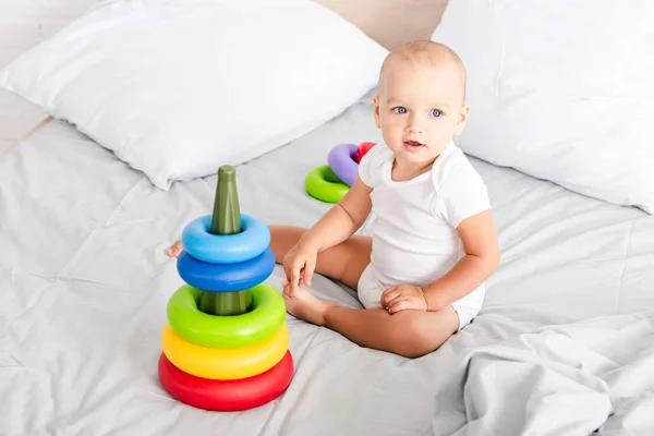 Criança descalça bonito em roupas brancas sentado na cama perto de pirâmide de brinquedos — Fotografia de Stock