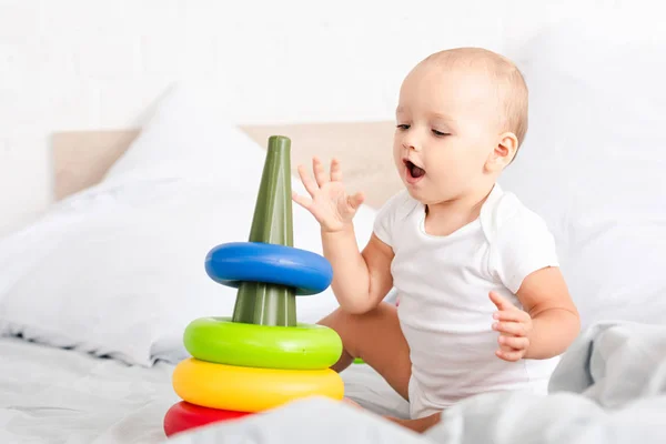 Cute little child in white clothes sitting on bed and playing with toy pyramid — Stock Photo