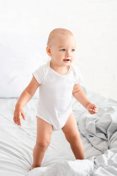 Niedliches kleines Kind in weißen Kleidern steht auf dem Bett, lächelt und schaut weg — Stockfoto