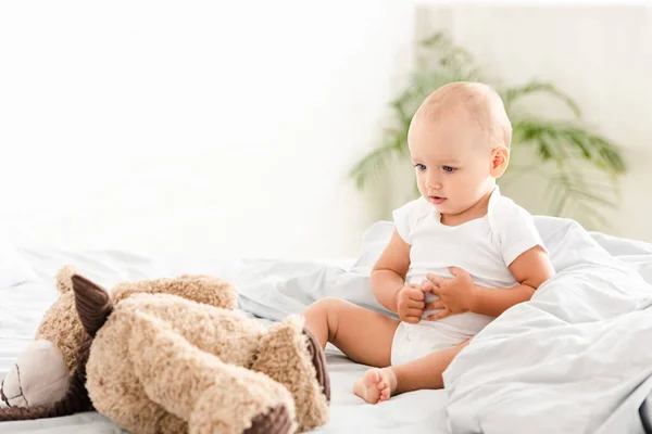 Маленький ребенок в белой одежде сидит на кровати и смотрит на плюшевого мишку — стоковое фото