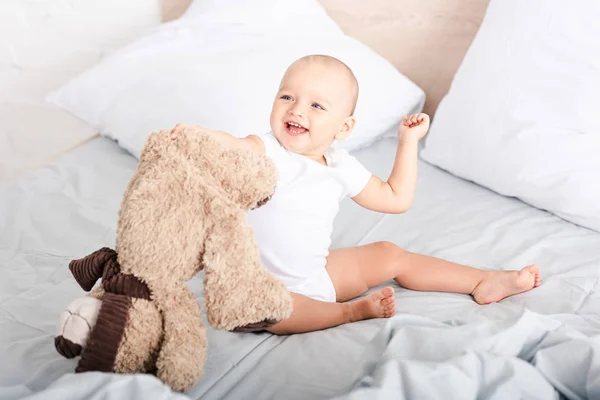 Смешной маленький ребенок в белой одежде сидит на кровати и смеется, держа плюшевого медведя — стоковое фото
