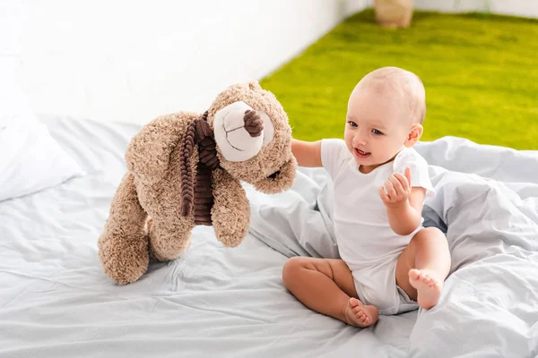 Смешной босоногий ребенок в белой одежде сидит на кровати и держит плюшевого медведя — стоковое фото