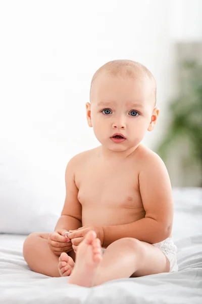 Cute barefoot child with blue eyes sitting on bed and looking at camera — Stock Photo