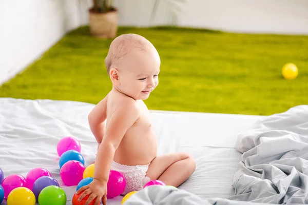 Funny little child sitting on bed among colored balls and smiling — Stock Photo