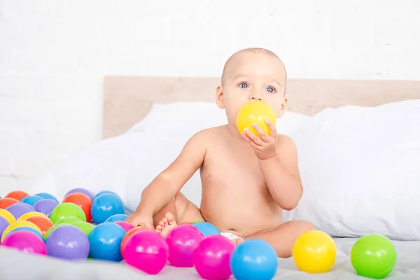 Lindo bebé sentado en la cama y tomando bola amarilla en la boca - foto de stock