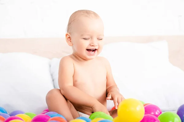 Cute little baby sitting on bed, playing with colored balls and laughing with closed eyes — Stock Photo
