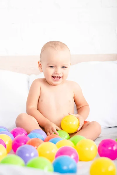 Niedliches kleines Baby sitzt auf dem Bett, spielt mit bunten Bällen und lacht — Stockfoto