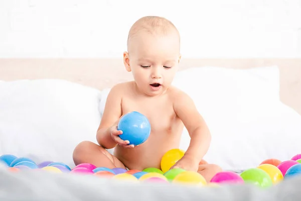 Niedliches kleines Baby sitzt auf dem Bett und spielt mit farbigen Kugeln — Stockfoto