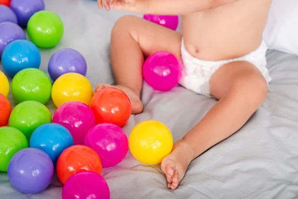 Vista parcial de pernas de bebê bonitos perto de pequenas bolas coloridas na cama branca — Fotografia de Stock