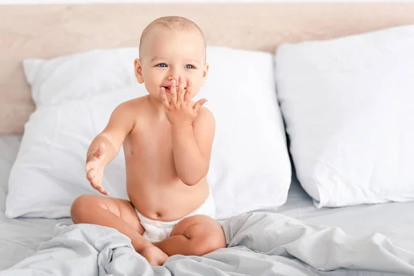 Adorable barefoot child in diaper smiling while sitting on white bedding — Stock Photo