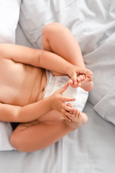 Vista parcial del pequeño bebé en pañal acostado en la espalda y tocando los pies con las manos - foto de stock