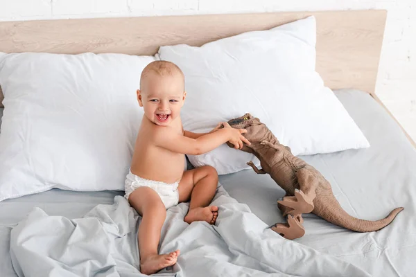 Criança pequena feliz sentada na cama, sorria e brincando com dinossauro de brinquedo marrom — Fotografia de Stock