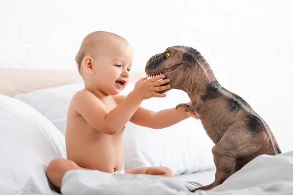 Маленький ребенок сидит на белой кровати и держит игрушечного динозавра обеими руками — стоковое фото