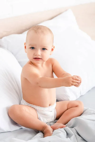 Cute blue-eyed child sitting on bed with white bedclothes and looking at camera — Stock Photo