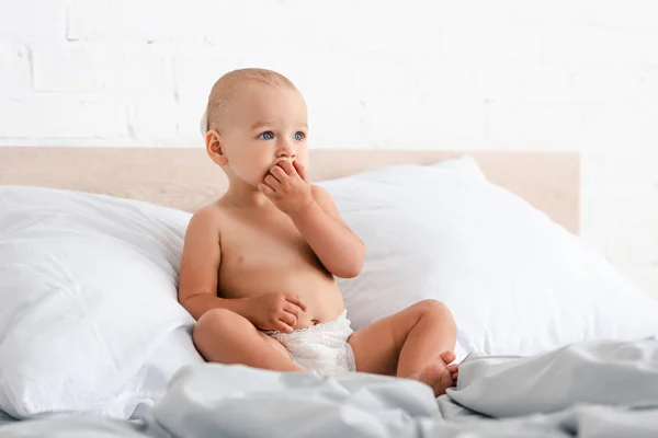 Barefoot little child sitting on bed and taking fingers into mouth — Stock Photo