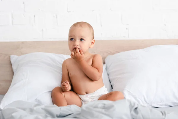 Adorable little child sitting on bed with pillows and taking fingers into mouth — Stock Photo