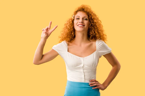 Sonriente mujer con el pelo rojo mostrando símbolo de victoria aislado en amarillo - foto de stock