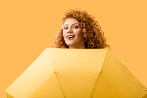 Улыбающаяся рыжая девушка позирует с зонтиком, изолированным на желтом — стоковое фото
