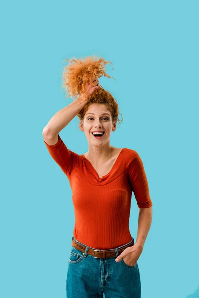 Jeune femme excitée tenant les cheveux bouclés rouges isolés sur bleu — Photo de stock