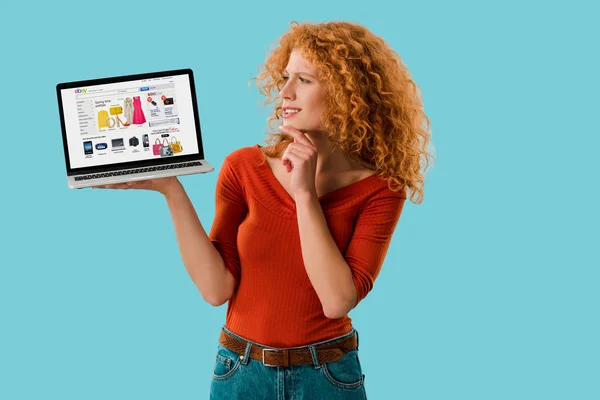 KYIV, UCRANIA - 16 de julio de 2019: pelirroja pensativa que sostiene el ordenador portátil con el sitio web de ebay, aislado en azul - foto de stock