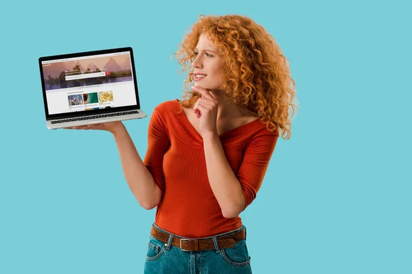 KYIV, UCRANIA - 16 de julio de 2019: pelirroja pensativa sosteniendo el ordenador portátil con obturador sitio web, aislado en azul - foto de stock