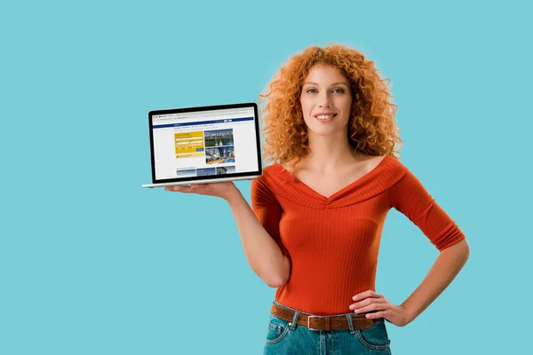 KYIV, UKRAINE - JULY 16, 2019: smiling redhead woman holding laptop with booking website, isolated on blue — Stock Photo