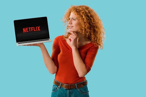 КИЕВ, УКРАИНА - 16 июля 2019 года: задумчивая рыжая женщина с ноутбуком netflix с веб-сайта, изолированные на синий — стоковое фото