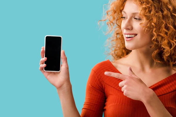 Chica pelirroja sonriente apuntando al teléfono inteligente con pantalla en blanco aislado en azul - foto de stock