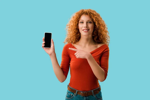 Mujer rizada sonriente apuntando al teléfono inteligente con pantalla en blanco aislado en azul - foto de stock
