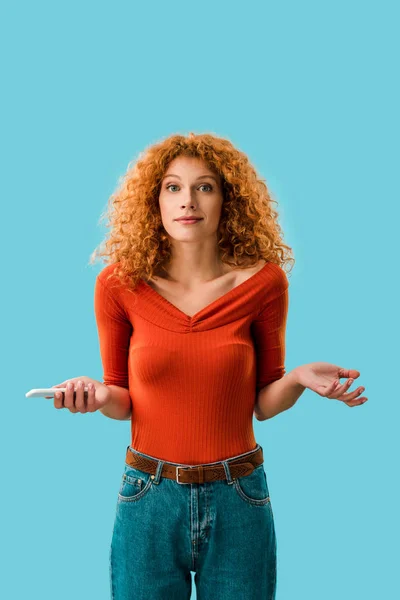 Confused redhead woman holding smartphone isolated on blue — Stock Photo