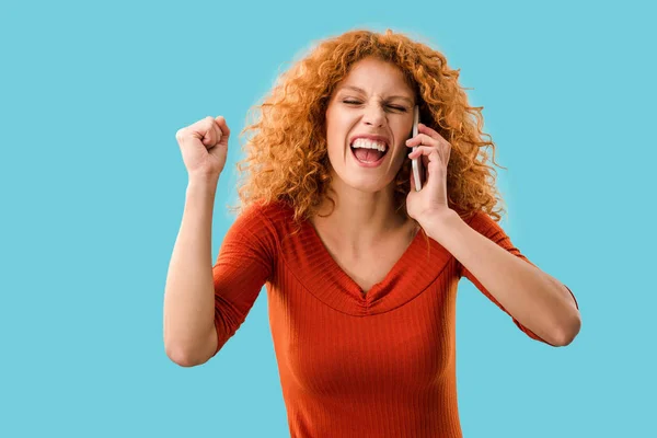 Excited curly redhead woman talking on smartphone isolated on blue — Stock Photo