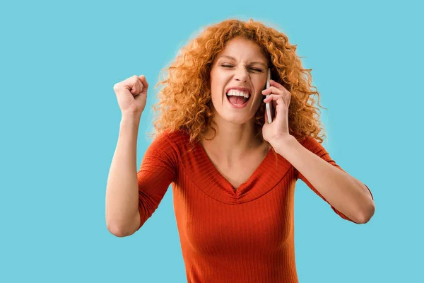 Excitada pelirroja rizada hablando en el teléfono inteligente aislado en azul - foto de stock