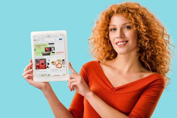 KYIV, UKRAINE - 16 JUILLET 2019 : femme rousse souriante tenant une tablette numérique avec application ebay, isolée sur bleu — Photo de stock