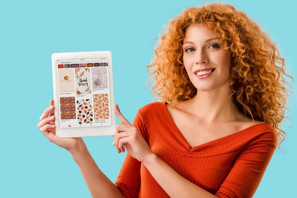 KYIV, UKRAINE - 16 JUILLET 2019 : femme rousse souriante tenant une tablette numérique avec application pinterest, isolée sur bleu — Photo de stock