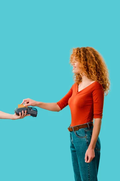 Rousse bouclée femme payant avec carte de crédit sur le terminal, isolé sur bleu — Photo de stock