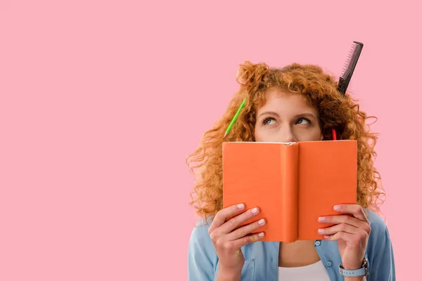 Redhead student with pencils in hair holding book isolated on pink — Stock Photo