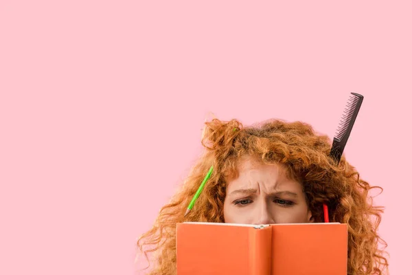 Концентрований рудий студент з олівцями у волоссі тримає книгу ізольовано на рожевому — стокове фото