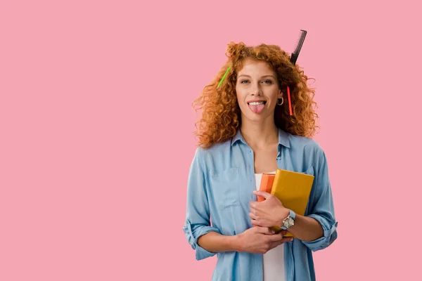 Pelirroja estudiante con lápices en el pelo sosteniendo libros y sacando la lengua aislado en rosa - foto de stock