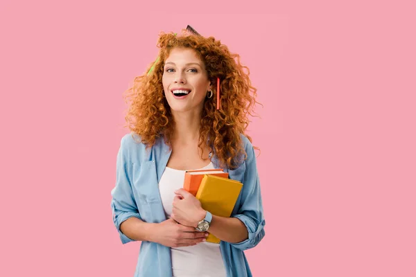 Happy redhead student with pencils in hair holding books isolated on pink — Stock Photo