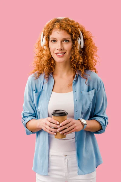 Rossa donna con carta tazza di caffè ascolto musica in cuffia, isolato su rosa — Foto stock
